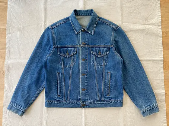 Front of a medium-wash denim trucker jacket laid flat on cream linen